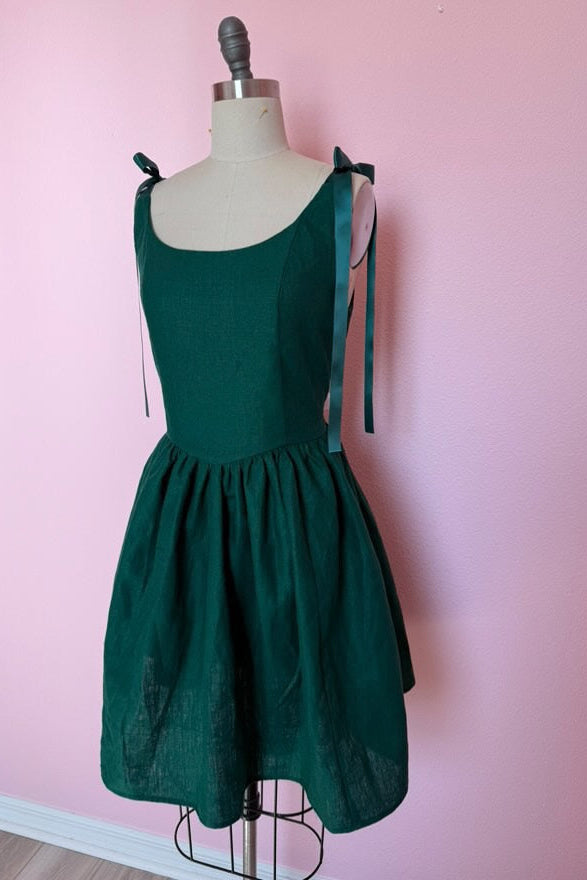 Custom made green linen dress with a scoop neck, gathered skirt, and satin ribbon tie straps. Shown on a mannequin with a pink background.