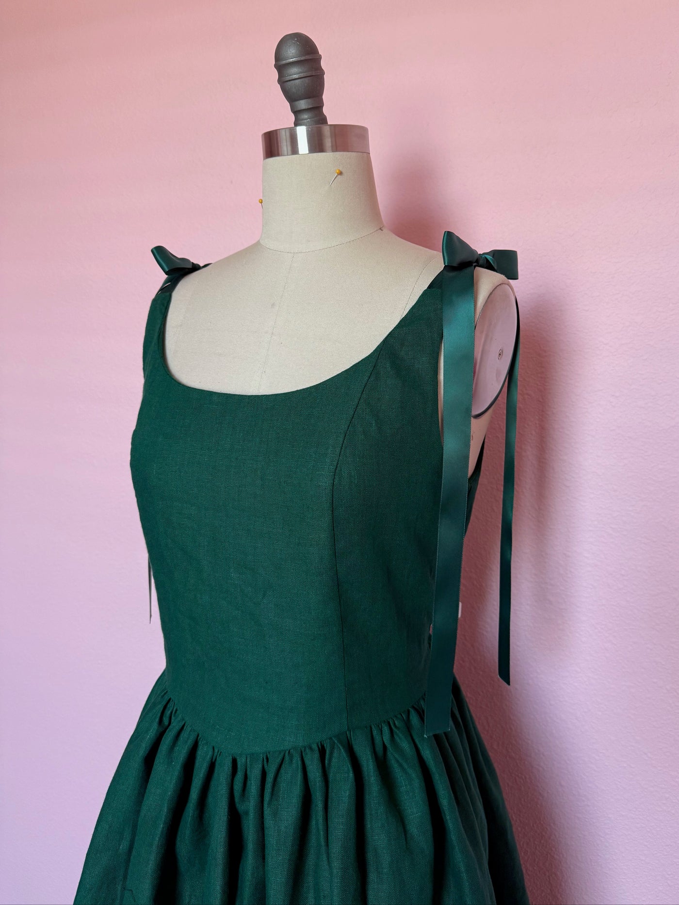 Green scoop neck dress with sati ribbon strap ties and gathered skirt on a mannequin against a pink wall.