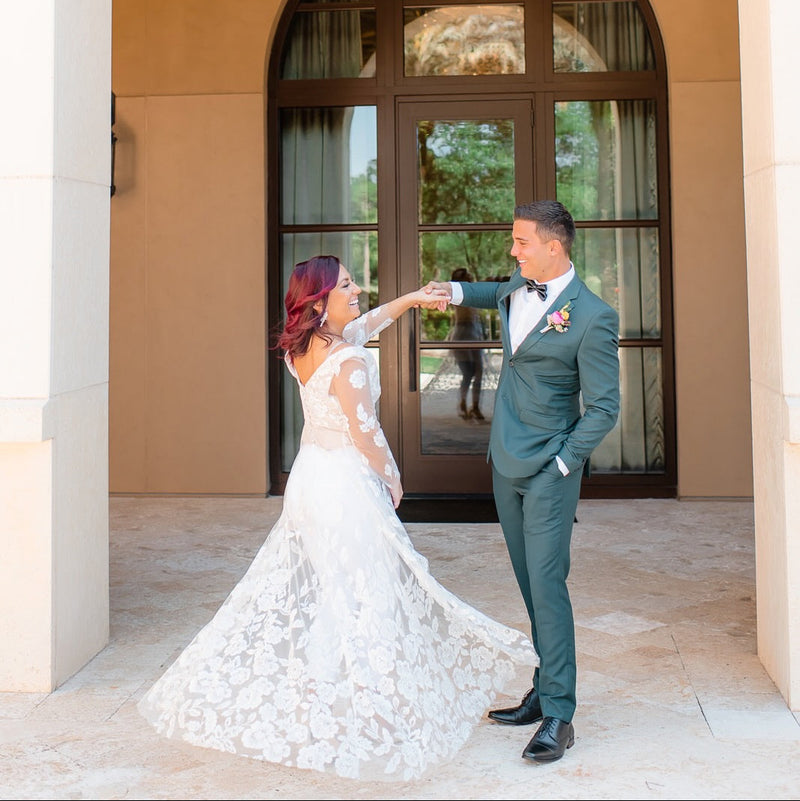 Couple in wedding attire standing under an archway with a building in the background. The woman is wearing a custom bridal bodice with long floral lace sleeves and a lace train.