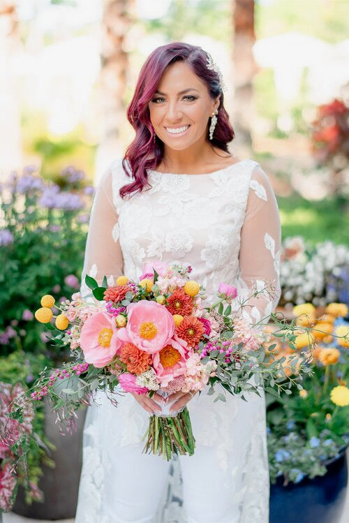 Bride holding a colorful bouquet of flowers in an outdoor garden setting. Bride is wearing a custom blush colored top with a white lace overlay, long white lace sleeves and train.
