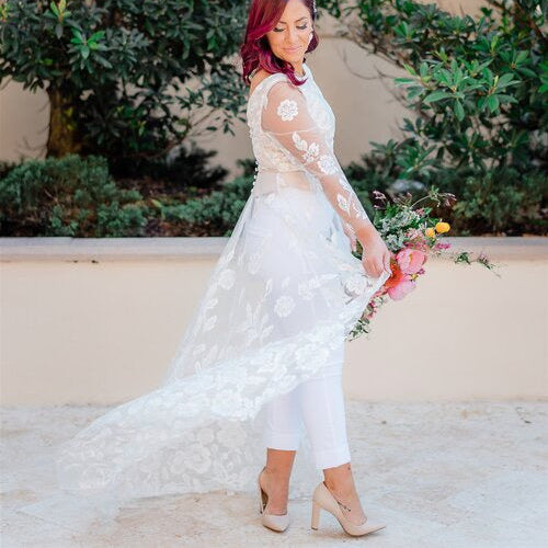 Woman is wearing a custom bridal top in blush satin with a floral lace overlay, long lace sleeves and train, paired with white pants and nude heels.