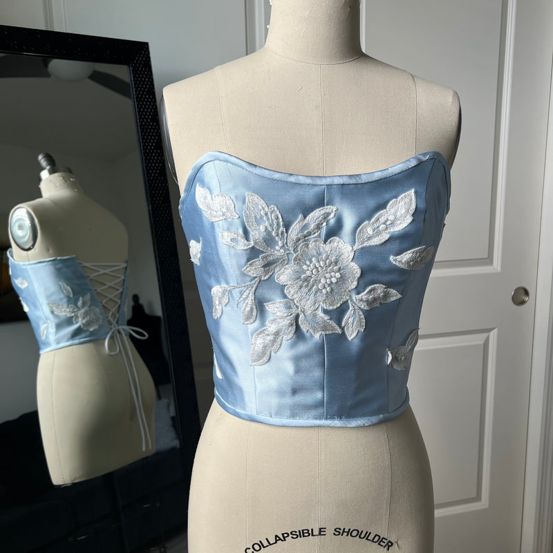 Baby blue satin twill  corset top with white floral lace applique on a mannequin with a mirror reflection in the background.